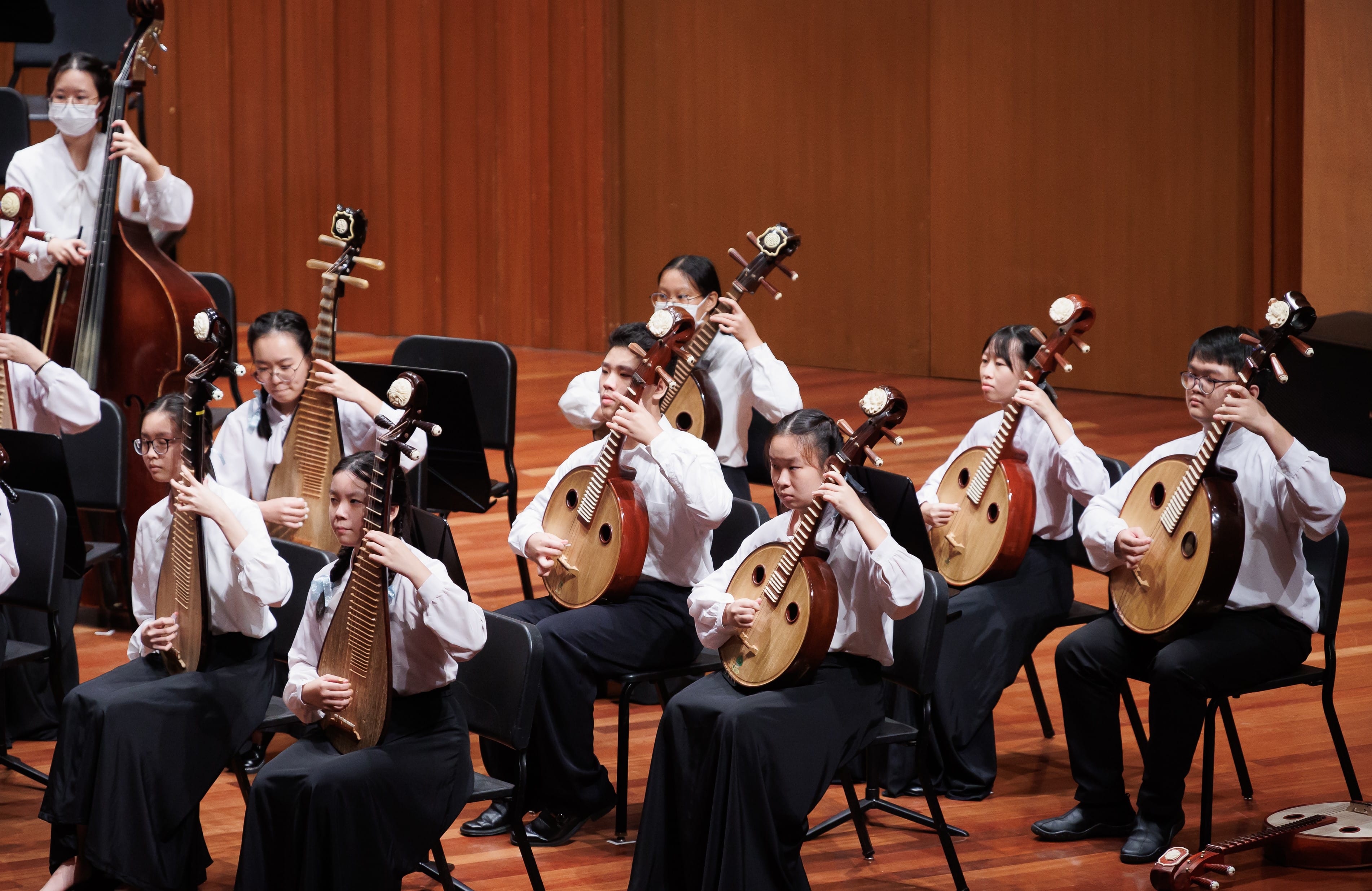 Young musicians in white shirts perform Chinese ruan and pipa instruments on a wooden concert stage.